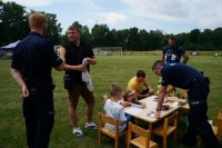 Dwaj policjanci przy stanowisku profilaktycznym prowadzą pogadanki z uczestnikami festynu. Dzieci siedzą przy stoliku. W lte murawa boiska i drzewa