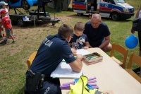 Policjant i chłopiec siedzą przy stoliku na trawie. Obok dziecka kuca mężczyzna. W tle widać karetkę pogotowia