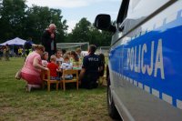 Na pierwszym planie policyjny radiowóz. Na drugim planie przy stoliku na trawie siedzą dzieci, przy nich kuca kobieta i policjant. Jeden mężczyzna stoi nad dziećmi, rozmawia z policjantem.