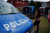 Na pierwszym planie policyjny radiowóz przy którym stoi kobieta z aparatem fotograficznym. W tle symulator dachowania