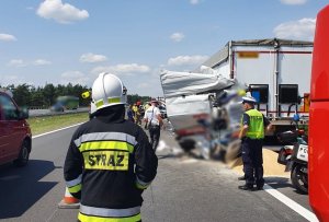 Miejsce wypadku drogowego. Na pierwszym planie stoi strażak, dalej dwóch poliocjantów. Na drugim planie tir uczestniczący w ywpadku stoi na poboczu