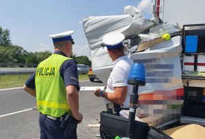 Miejsce wypadku drogowego. Na pierwszym planie stoi dwóch policjantów, na drugim planie na poboczu stoi tir uczestniczący w wypadku