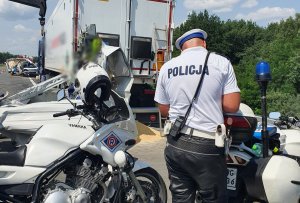 Miejsce wypadku drogowego,na pierwszym planie przy motocyklu stoi policjant sporzadza dokumentację. Na drugim planie na poboczu stoi tir uczestniczący w wypadku