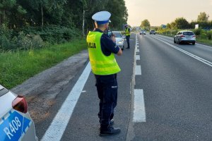 Policjant ruchu drogowego w odblaskowej zielonej kamizelce stoi tyłem z miernikiem prędkości. W tle jezdnia i samochody.