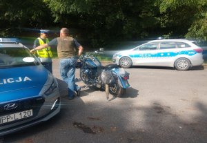 Policjant ruchu drogowego w trakcie działań, kontroluje kierowcę motocykla. Obok stoją dwa radiowozy i motocykl. W tle widać drzewa