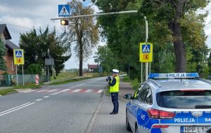 BNa pierwszym planie w prawym dolnym rogu widać policyjny radiowóz. Na drugim planie w rejonie przejścia dla pieszych stoi policjantka ruchu drogowego