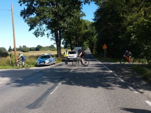 Na pierwszym planie ulica, na drugim planie policjant stoi przy radiowozie, wstrzymuje ruch samochodów, przez jezdnię przejeżdżają rowerzyści