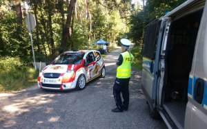 Policjantka ruchu drogowego w odblaskowej kamizelce stoi przy radiowozie. W tle widać samochód rajdowy jadący drogą biegnącą przez las