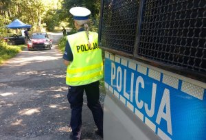 Policjantka ruchu drogowego w odblaskowej kamizelce stoi przy radiowozie. W tle widać samochód rajdowy jadący drogą biegnącą przez las