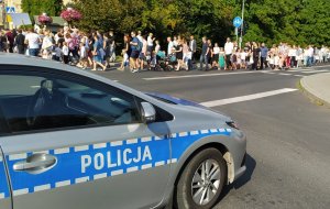 Na pierwszym stoi radiowóz policyjny. W tle korowód dożynkowy idzie ulicą. Na dalszym planie widać drzewa