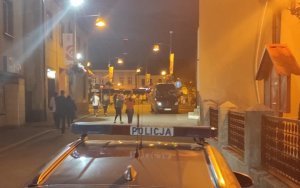 Zdjęcie wykonane nocą, przy oświetleniu latarni. Na drodze dojazdowej do Rynku stoi radiowóz policyjny. W tle na Rynku widać uczestników imprezy
