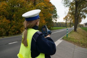 Policjantka ruchu drogowego w odblaskowej kamizelce stoi na poboczu jezdni mierzy prędkość pojazdów. W tle widać jezdnię i drzewa