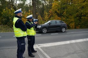 Policjanci ruchu drogowego w odblaskowych kamizelkacg stoją na poboczu jezdni. Policjantka mierzy prędkość pojazdów. W tle widać jezdnię i drzewa oraz przemieszczający się czarny samochód osobowy