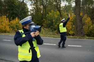 Na pierwszym planie policjantka ruchu drogowego w odblaskowej kamizelce stoi na poboczu jezdni i mierzy prędkość pojazdów. Na drugim planie stoi na jezdni z uniesioną do góry ręką zatrzymując pojazd do kontroli. W tle widać drzewa