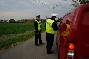Policjanci ruchu drogowego w odblaskowych kamizelkach stoją przy zatrzymanym do kontroli czerwonym samochodzie. W tle widać pole i zabudowania