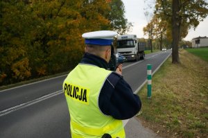 Policjant ruchu drogowego w odblaskowej kamizelce stoi na poboczu jezdni i mierzy prędkość pojazdów. W tle widać jezdnię którą przemieszcza się samochód ciężarowy i drzewa