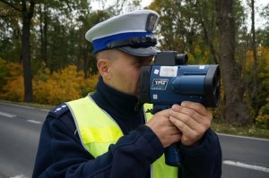 Policjant ruchu drogowego w odblaskowej kamizelce mierzy prędkosć pojazdów. W tle widać jezdnię i drzewa