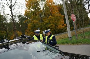 Policjanci ruchu drogowego w odblaskowych kamizelkach stoją przy zatrzymanym do kontroli samochodzie. W tle widać jezdnię, drzewa i znak drogowy STOP