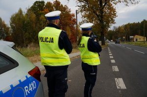 Policjanci ruchu drogowego w odblaskowych kamizelkach stoją na poboczu jezdni przy radiowozie. Policjantka mierzy prędkość pojazdów. W tle widać jezdnię, jadące samochody i drzewa