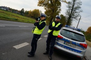 Policjanci ruchu drogowego w odblaskowych kamizelkach stoją na poboczu jezdni przy radiowozie. Policjantka mierzy prędkość pojazdów. W tle widać jezdnię i drzewa