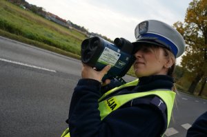 Policjantka ruchu drogowego w odblaskowej kamizelce mierzy prędkość pojazdów. W tle widać jezdnię i drzewa