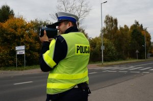 Policjant ruchu drogowego w odblaskowej kamizelce stoi na poboczu jezdni i mierzy prędkosć pojazdów. W tle widać jezdnię, przejśćie dla pieszych i drzewa