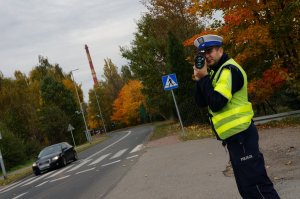 Policjant ruchu drogowego w odblaskowej kamizelce stoi na poboczu jezdni i mierzy prędkość pojazdów. W tle widać jezdnię po której jedzie czarny samochód, przejście dla pieszych i drzewa