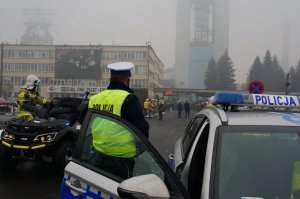 Policjant ruchu drogowego stoi przy radiowozie. W tle widać kopalnie i osoby uczestniczace w obchodach rocznicy