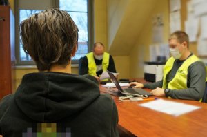 Zatrzymany 16-latek w trakcie przesłuchania w pomieszczeniu Komendy Powiatowej Policji w Bieruniu. Chłopiec na pierwszym planie siedzi tyłem. W tle dwaj policjanci dochodzeniowo-śledczy w odblaskowych kamizelkach siedzą za biurkami.