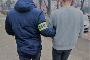 Nieumundurowany policjant, który na ramieniu ma opaskę z napisem POLICJA, trzyma pod rękę zatrzymanego mężczyznę