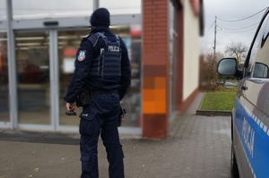 Umundurowany policjant stoi tyłem, na wprost wejścia do sklepu. W prawej części zdjęcia widać fragment radiowozu