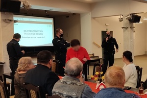 Na pierwszym planie osoby starsze siedzą za stołem. Na drugim planie stoją policjanci, a za nimi znajduje się ekran z prezentacją