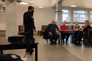 Na pierwszym planie w lewej części zdjęcia stoi policjant, który trzyma przy ustach mikrofon. Na drugim planie seniorzy siedzą za stołem.