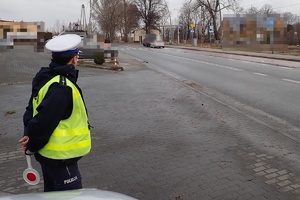 Policjant ruchu drogowego w odblaskowej kamizelce stoi w pobliżu jezdni i obserwuje skrzyżowanie i przejście dla pieszych. Policjant trzyma z tyłu tarczę STOP