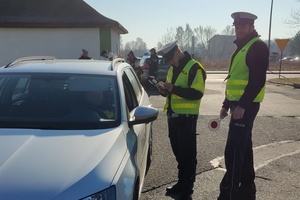 Policjanci ruchu drogowego stoją przy kontrolowanym samochodzie. W tle widać budynek i ulicę