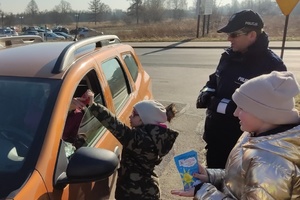 Policjant i dzieci stoją przy pomarańczowym samochodzie. W tle widać ulicę