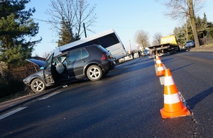 Miejsce wypadku. Na pierwszym planie jezdnia i rozstawione pachołki drogowe. Dalej widać dwa samochody w przydrożnym rowie a za nimi pojazdy służb ratowniczych