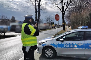 Policjant ruchu drogowego stoi na poboczu jezdni i prowadzi pomiar prędkości pojazdów. W tle widać jezdnię, przejście dla pieszych, na poboczu znak drogowy STOP