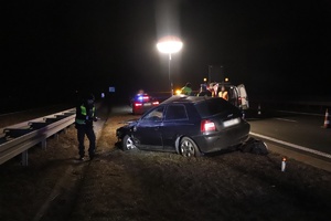 Miejsce wypadku. Zdjęcie wykonane w porze nocnej. Rozbity samochód stoi na pasie zieleni przy barierach. Policjant przy samochodzie robi oględziny. W tle widać oświecającą miejsce zdarzenia dużą lampę