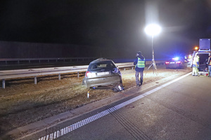 Miejsce wypadku. Zdjęcie wykonane w porze nocnej. Rozbity samochód stoi na pasie zieleni przy barierach. Policjant przy samochodzie robi oględziny. W tle widać oświecającą miejsce zdarzenia dużą lampę