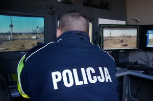 Policjant ruchu drogowego siedzi tyłem, przed nim monitory, na których obserwuje przejazd kolejowy. Na plecach, na mundurze policjanta widać napis: POLICJA