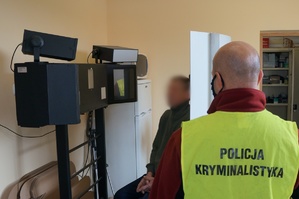Na pierwszym planie tyłem stoi nieumundurowany policjant wydziału kryminalnego w odblaskowej kamizelce z napisem na plecach: POLICJA KRYMINALISTYKA. W tle widać podejrzanego, który siedzi na urządzeniu do wykonywania zdjęć sygnalitycznych.