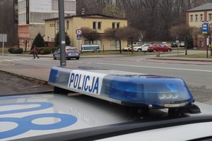 Listwa sygnalizacyjna radiowozu z napisem POLICJA na tle jezdni i przejścia dla pieszych, przez które przechodzi pieszy. Przed przejściem stoi policyjny radiowóz, ustępuje pierwszeństwa pieszemu.