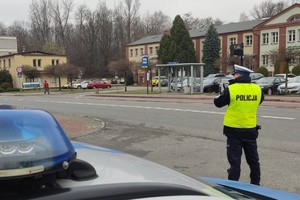 Po prawej stronie policjant ruchu drogowego stoi tyłem w odblaskowej kamizelce z napisem na plecach: POLICJA. W tle widać jezdnię, przejście dla pieszych, budynki i drzewa. Na pierwszym planie widać fragment radiowozu.