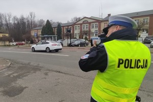 Na pierwszym planie po prawej stronie policjant ruchu drogowego stoi tyłem w odblaskowej kamizelce z napisem na plecach: POLICJA. W tle widać jezdnię, przejście dla pieszych, budynki i drzewa. Jezdnią jedzie samochód.