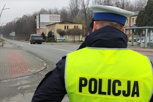 Na pierwszym planie po prawej stronie policjant ruchu drogowego stoi tyłem w odblaskowej kamizelce z napisem na plecach: POLICJA. W tle widać jezdnię, przejście dla pieszych, budynki i drzewa. Do przejścia dojeżdża ciężarowy samochód.