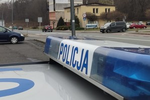 Na pierwszym planie listwa sygnalizacyjna radiowozu z napisem POLICJA. W tle widać jezdnię i przejście dla pieszych.