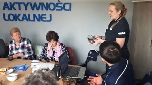 W prawej części zdjęcia dwoje policjantów prowadzących prelekcję, policjantka stoi, policjant siedzi. W lewej części widać starsze osoby siedzące za stołem. Na ścianie znajduje się napis: CENTRUM AKTYWNOŚCI LOKALNEJ