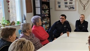 Prelekcja u seniorów. Osoby starsze siedzą za stołem, obok siedzą policjanci, których widać na drugim planie. W tle widać okno, czarne meble i ozdoby na ścianie