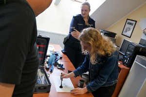 Praktykantka w pokoju technika kryminalistyki, trzyma w ręku pędzelek, którym porusza po kartce ze śladami daktyloskopijnymi. Na drugim planie stoi policjantka w mundurze